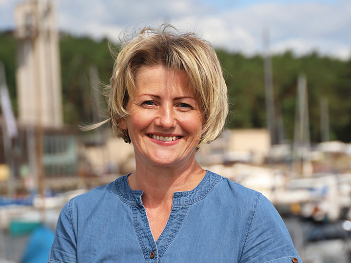 Natural Pharmaceuticals „CalciumControl™“ Šypsanti vidutinio amžiaus moteris trumpais šviesiais plaukais prie uosto su laivais ir medžiais fone.