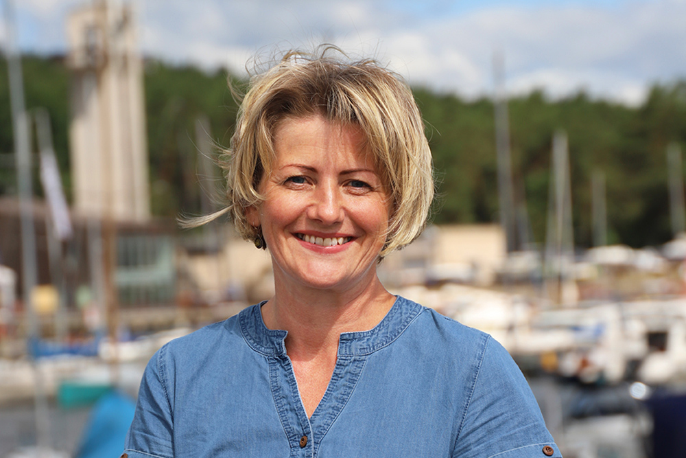 Natural Pharmaceuticals „CalciumControl™“ Šypsanti vidutinio amžiaus moteris trumpais blondiniais plaukais ir mėlyna palaidine prie uosto ir laivų fono.