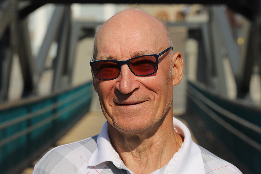 Natural Pharmaceuticals „CalciumControl™“ Vyresnio amžiaus vyras su akiniais nuo saulės ir balta palaidine stovi lauke ant tilto.