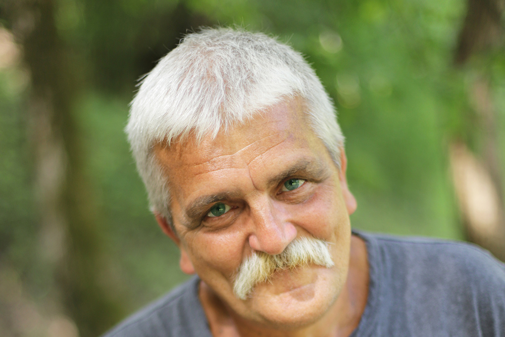 Natural Pharmaceuticals „CalciumControl™“ Vyresnio amžiaus vyras su baltais plaukais ir ūsais žvelgia į kamerą gamtos fone.